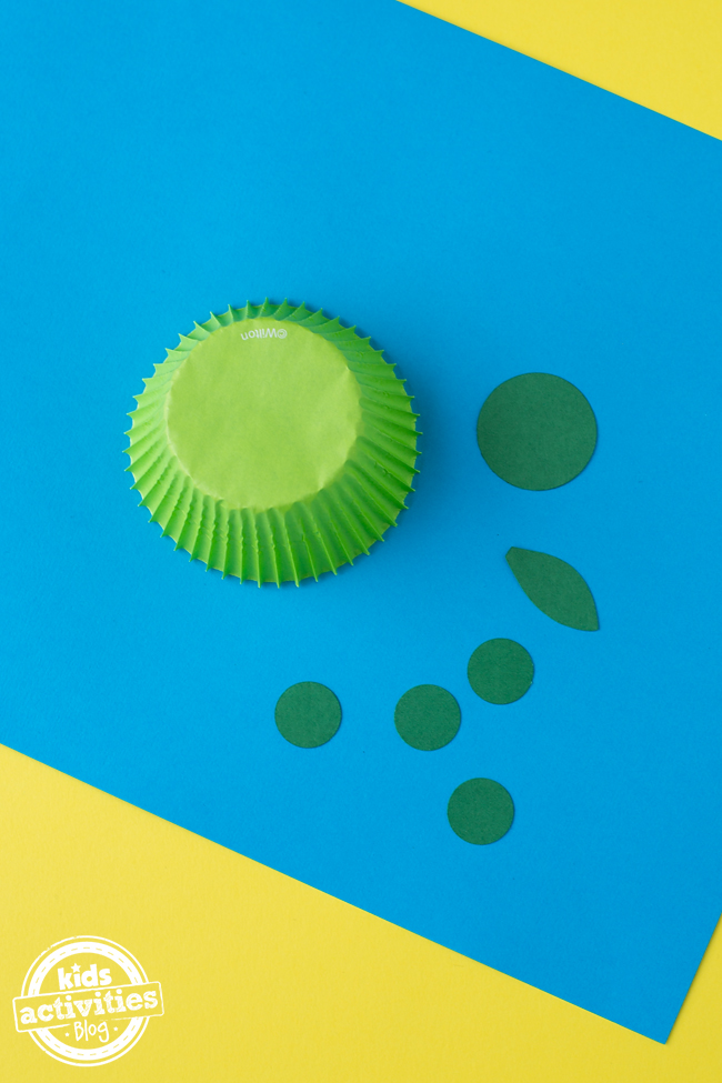 Cupcake Liner Turtle Craft - Étape 2 - découpez 4 cercles de papier de construction vert, une queue ovale et un cercle pour la tête sur du papier de construction bleu avec une doublure de cupcake verte contre une table jaune - blog d'activités pour enfants