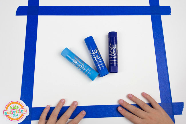 Winter Forest Preschool Art - blog d'activités pour enfants - étape 1 - morceau de papier scotché avec du ruban de peintre bleu avec des bâtons de peinture bleu foncé, bleu et bleu clair