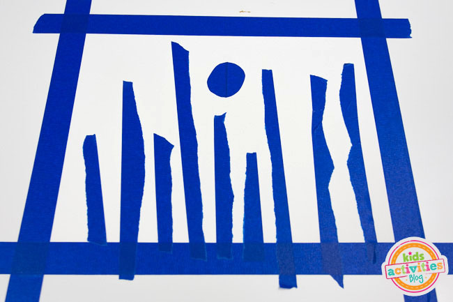 Winter Forest Preschool Art - blog d'activités pour enfants Étape 2 - ruban de peintre bleu déchiré en deux et mis sur du papier en cercle pour faire une lune et verticalement pour faire des arbres pour la peinture de résistance.
