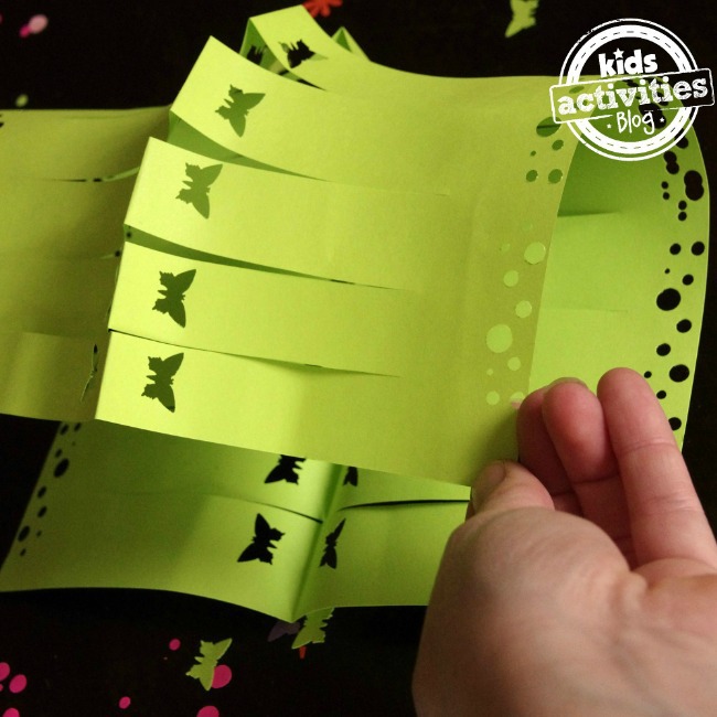 Étape 4 - Comment utiliser un perforateur pour créer des papillons et des petits et grands points sur une lanterne en papier verte qui est pliée par une petite main.