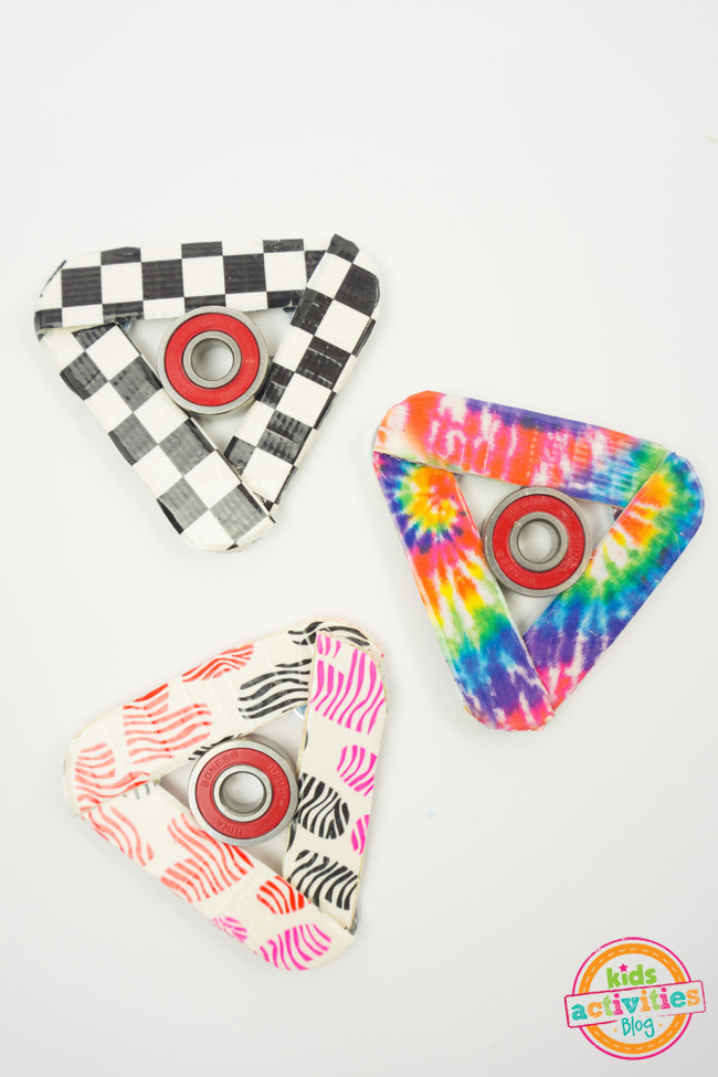 Comment faire de l'artisanat Fidget Spinner - trois spinners faits maison terminés montrés - un avec du ruban adhésif en damier, un décoré avec du ruban adhésif arc-en-ciel et un avec des coeurs multicolores