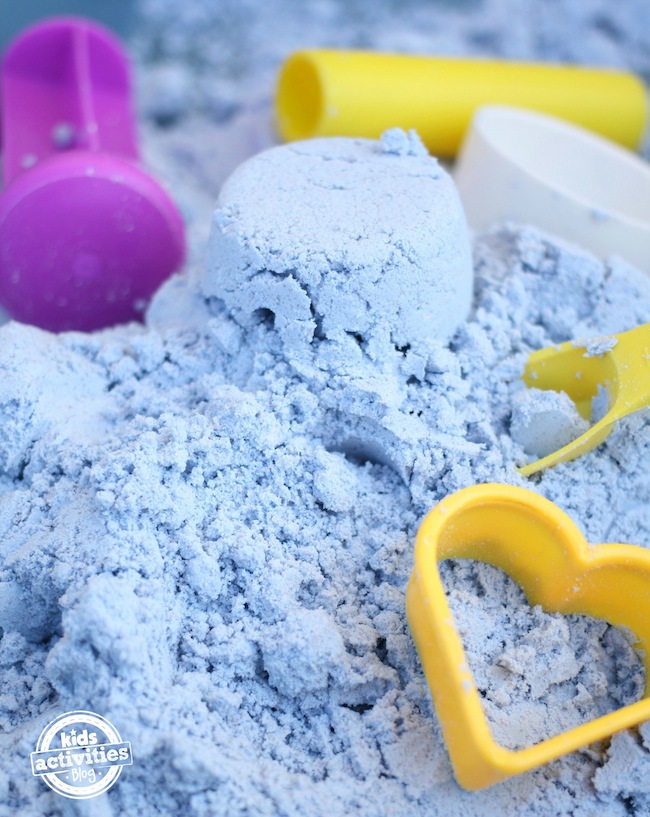 Pâte nuageuse sans danger pour les tout-petits dans un tas bleu avec des jouets en plastique, des pelles et un moule en forme de cœur.