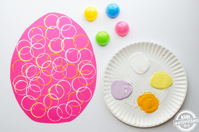 étape 4 - artisanat d'oeufs de pâques préscolaire - cercles fabriqués à partir d'un tampon d'oeuf de pâques illustré partout sur un oeuf en papier de construction avec une assiette en papier de peinture à côté