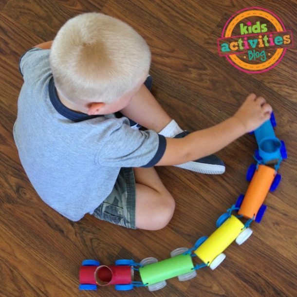 Des activités sans écran comme ce train de rouleaux de papier toilette avec lequel ce petit garçon joue ! Petit garçon aux cheveux blonds et une chemise grise et un pantalon camo et des chaussettes blanches avec des chaussures à enfiler noires assis sur un plancher en bois jouant avec un train de rouleau de papier toilette. Il a la voiture principale, 3 voitures régulières et le caboose. Le train est bleu, orange, jaune, vert et rouge, relié par une ficelle bleue et a des roues bleues et claires et blanches.