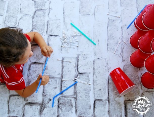 des activités sans écran comme ces jeux d'empilage de gobelets ou de ramasser des jeux de paille. Un petit garçon aux cheveux bruns dans une chemise rouge et blanche souffle des Q-Tips à travers des pailles bleues et vertes sur des tasses solo rouges pour les faire tomber. Il y a 9 tasses solo rouges empilées et 1 posée sur le sol.