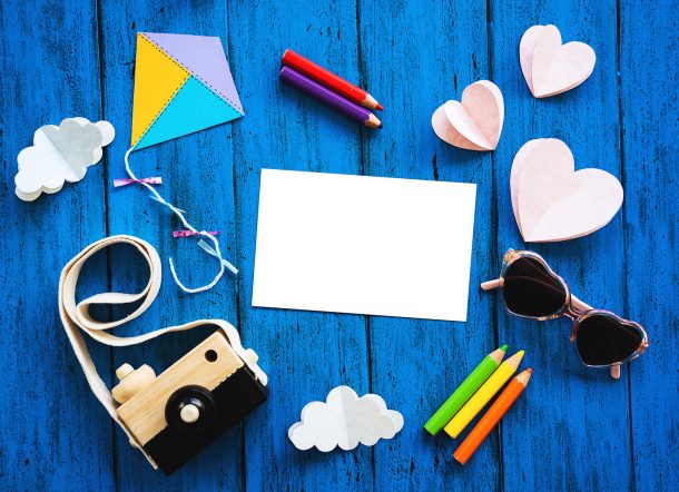 Écran des activités gratuites pour les enfants avec du papier, des fournitures de coloriage, comme un crayon de couleur rouge, violet, vert, jaune et orange, un cerf-volant en papier jaune, bleu et violet avec une longue queue avec des rubans violets, des nuages de papier 3d qui sont blancs , et coeurs roses en papier 3d. Il y a un appareil photo beige et noir et des lunettes en forme de cœur roses et violettes sur un fond en bois bleu.
