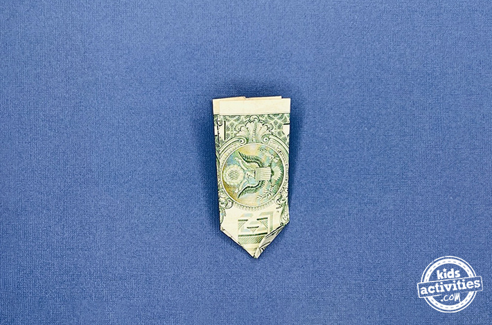 Pantalon origami en forme de billet d'un dollar - Étape 5 - Pliez le coin inférieur gauche vers le haut, puis dépliez-le pour créer un pli. Utilisez ce pli pour rentrer soigneusement le coin entre les deux couches du billet. Répétez avec le coin inférieur droit.
