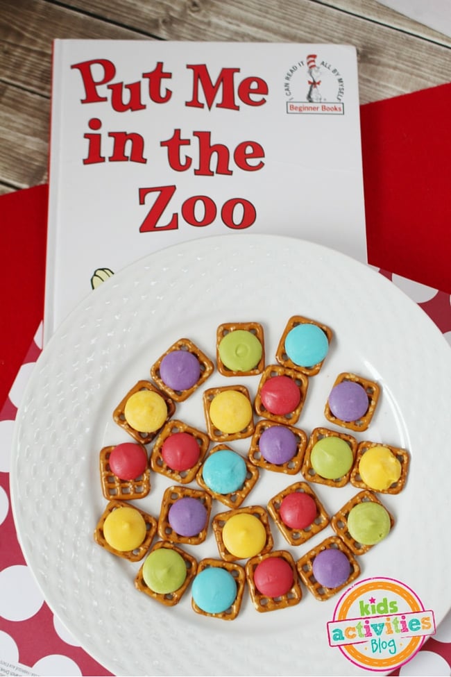 L'artisanat comestible m'a mis dans la collation du zoo avec des bretzels carrés et des bonbons rouges, bleus, violets, verts et jaunes fondants sur une assiette blanche avec le livre Mettez-moi dans le zoo sur fond rouge et fond de bois