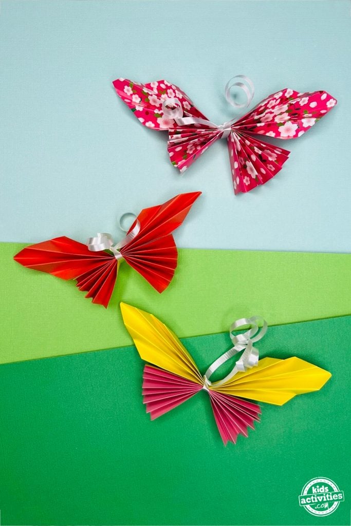 3 créations papillons en papier finies avec différents motifs et couleurs sur un fond coloré.  - Blog d'activités pour enfants