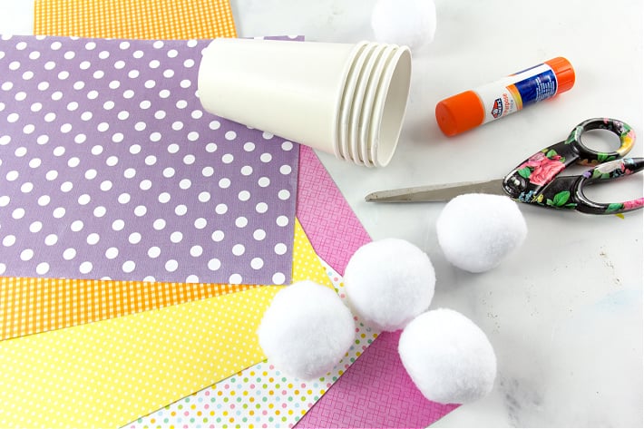 Les fournitures nécessaires pour fabriquer des gobelets à collations en forme de fesses de lapin pour Pâques comprennent des gobelets en papier, des pompons et du papier de scrapbooking.