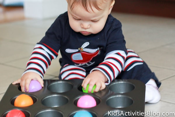 Muffin Tins and Ball Activity - jeune bébé jouant avec un moule à muffins en métal et des boules en plastique arc-en-ciel sur un sol carrelé - blog d'activités pour enfants