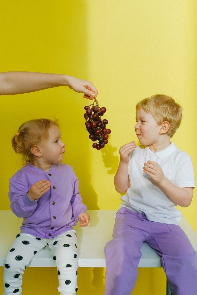 enfants mangeant des raisins assis sur un banc devant un mur jaune