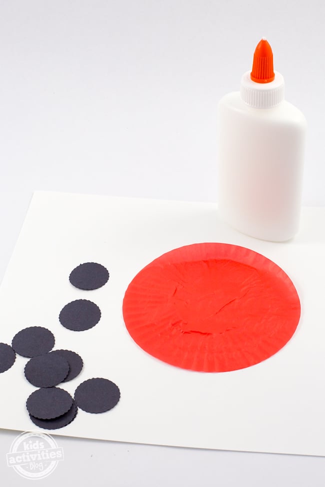 Étape 1 - Cupcake Liner Ladybug craft - colle blanche, doublure de cupcake rouge aplatie sur du papier blanc avec des cercles de papier de construction noir.