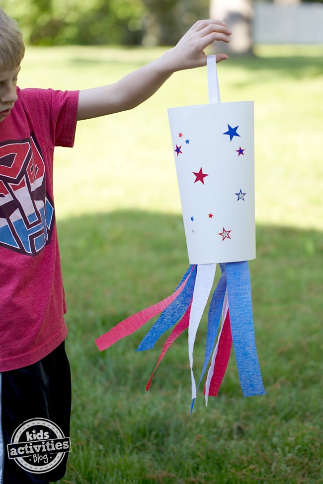 Étape 2 - Comment fabriquer une manche à air patriotique en papier - enfant tenant la manche à air en forme de cylindre avec des étoiles et des banderoles rouges, blanches et bleues
