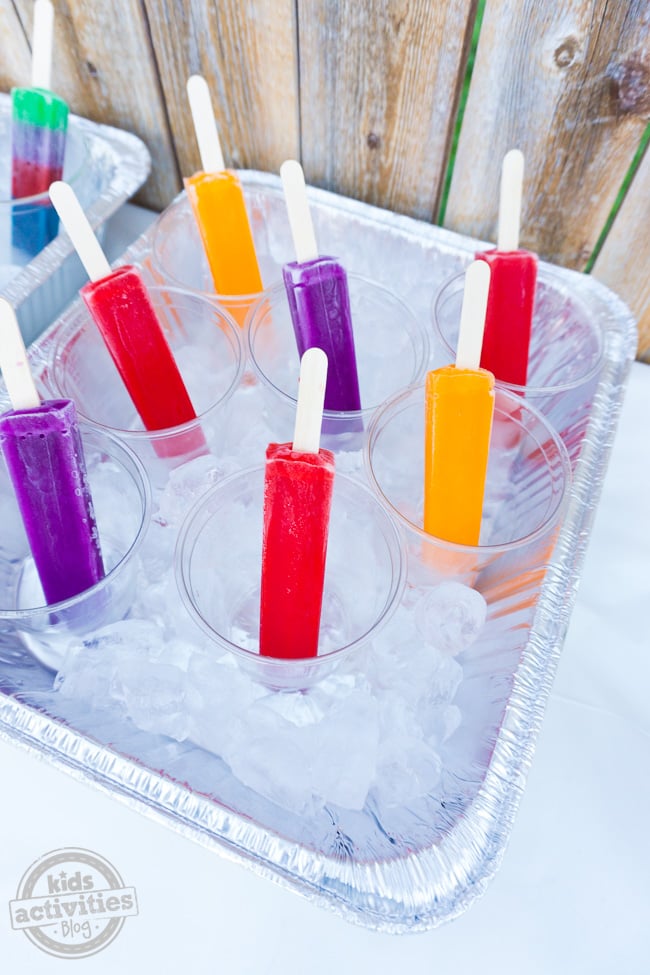 Summer Party Popsicle Bar illustré contre une clôture avec une table contenant de la glace et des glaces dans des tasses que les enfants peuvent saisir