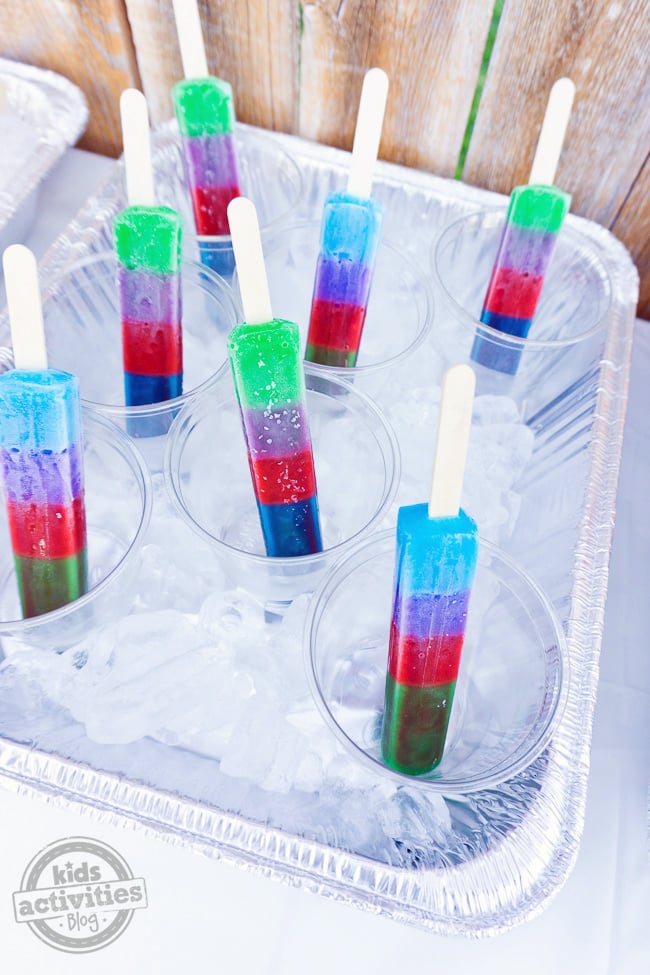 Summer Party Popsicle Bar avec des glaces à rayures vertes, rouges, violettes et bleues servies dans des tasses glacées dans une casserole de glace