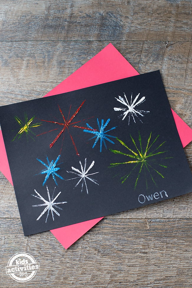 Artisanat de feux d'artifice - papier noir avec feux d'artifice de peinture rouge, jaune, blanc et bleu avec des paillettes sur fond rouge et bois - blog d'activités pour enfants