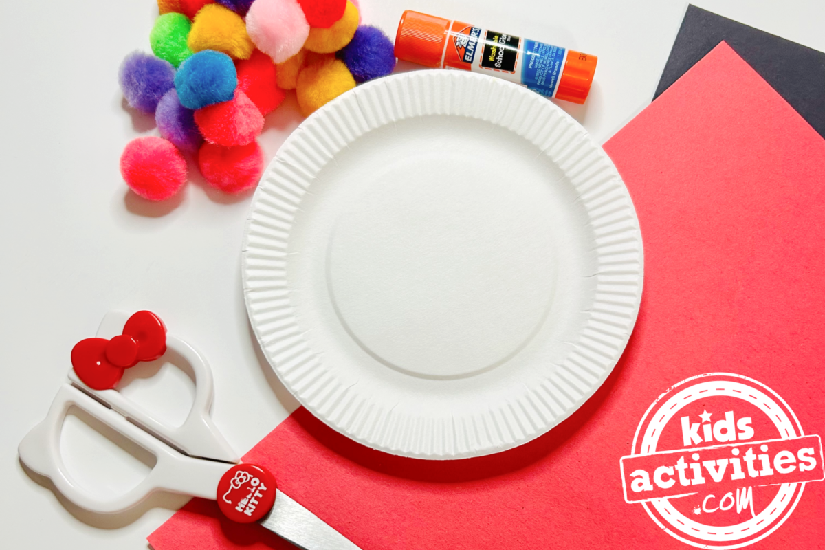 Fournitures nécessaires pour fabriquer une machine à boules de gomme avec une assiette en carton : du papier de construction rouge et noir, une mini assiette en carton, des ciseaux, des pompons artisanaux et de la colle - Blog d'activités pour enfants