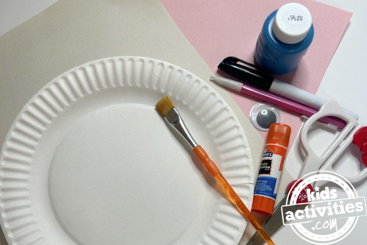 Fournitures nécessaires pour le bricolage d'une assiette en carton de narval : une assiette en carton, du papier de construction blanc et rose, de la peinture bleue, un œil mobile, un crayon, un feutre, des ciseaux et un pinceau - Blog d'activités pour enfants