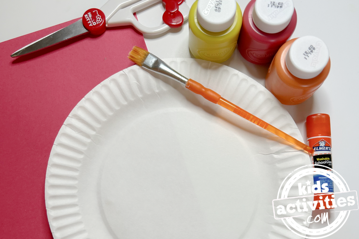 Fournitures nécessaires pour la fabrication d'une assiette à pizza en papier : une assiette en carton, du papier de construction rouge, de la peinture jaune, rouge et orange, des ciseaux, un pinceau et de la colle - Blog d'activités pour enfants