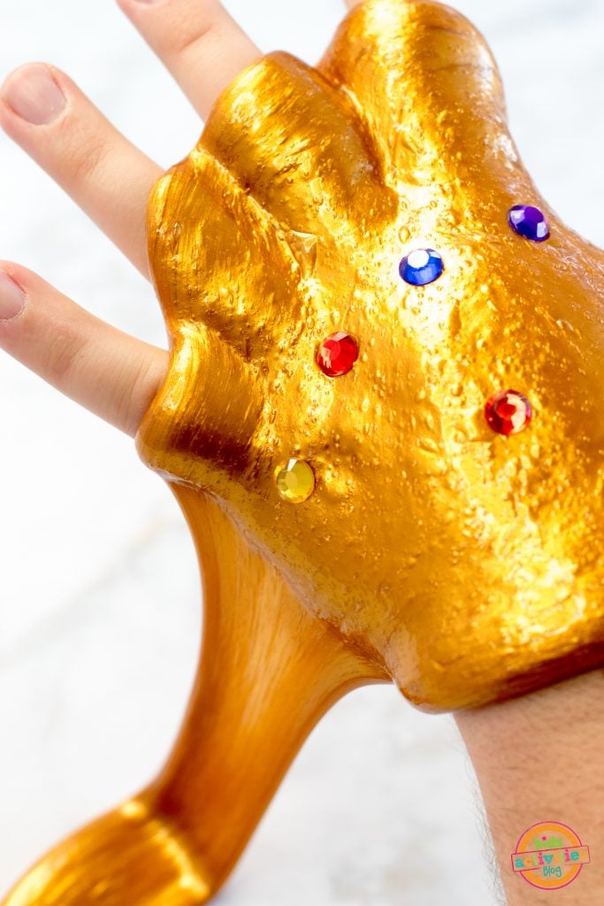 Slime Infinity Gauntlet fait maison, doré et extensible, avec des pierres précieuses rouges, bleues, violettes et jaunes sur la main de quelqu'un - Blog d'activités pour enfants