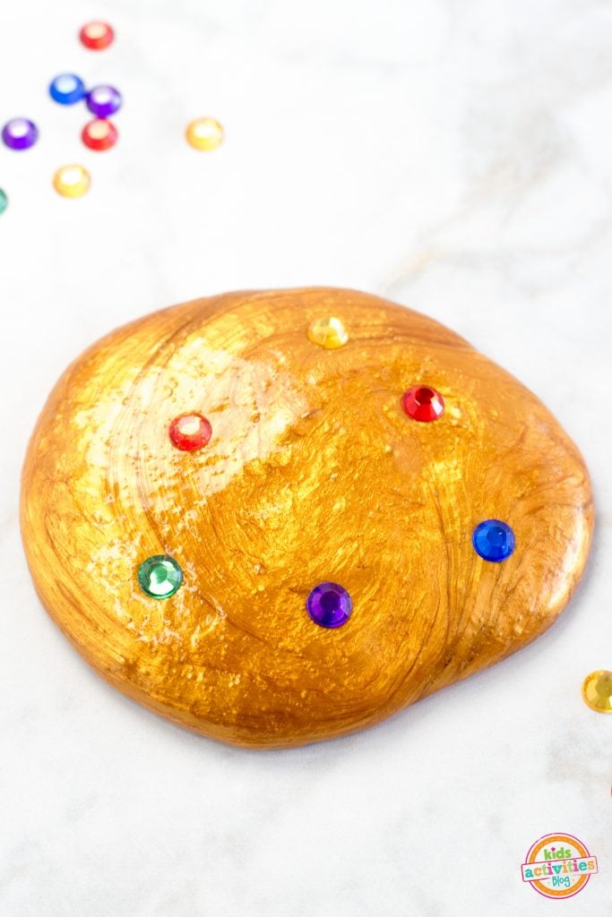 Slime Infinity Gauntlet fait maison - doré avec des pierres précieuses rouges, orange, jaunes, vertes, violettes et bleues - basé sur Marvels Avengers sur un fond blanc avec des pierres précieuses en vrac - Blog d'activités pour enfants