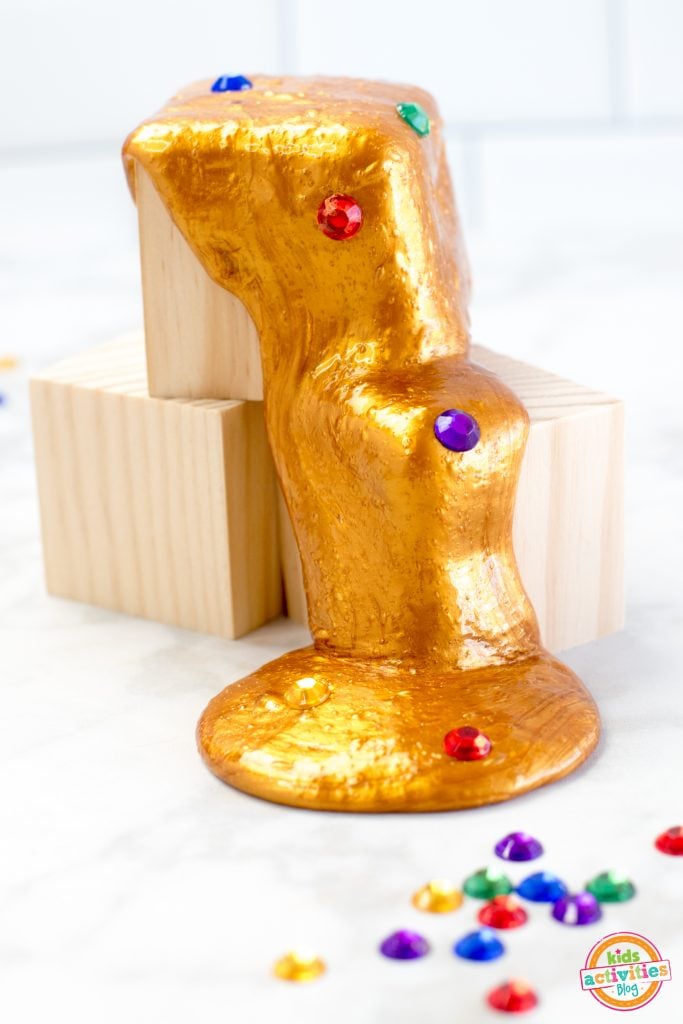 Slime fait maison de type gantelet de l'infini qui suinte de blocs de bois empilés sur un fond blanc avec des pierres précieuses rouges, bleues, vertes, jaunes et violettes dispersées - blog d'activités pour enfants