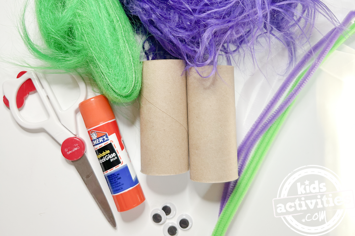 Fournitures nécessaires pour fabriquer un monstre en peluche : deux rouleaux de papier toilette, du tissu en fourrure violet et vert, des yeux mobiles, de la colle, des ciseaux et des cure-pipes