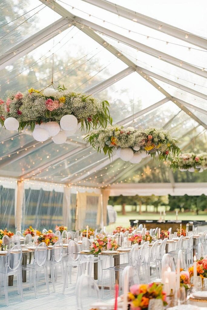 Tente transparente avec lustres floraux pour réception de mariage dans la cour.
